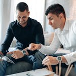Two businessmen in casual clothing discuss a project on a tablet indoors.